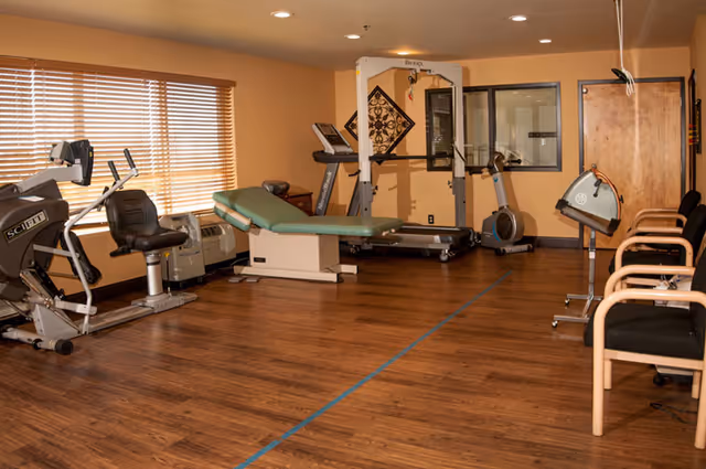 A fitness room with exercise equipment including a recumbent bike, treadmill, rowing machine, and a weight machine. There is a padded examination or therapy table in the center, several chairs lined up against the wall, and wooden blinds covering the windows. The room has wood flooring and beige walls.