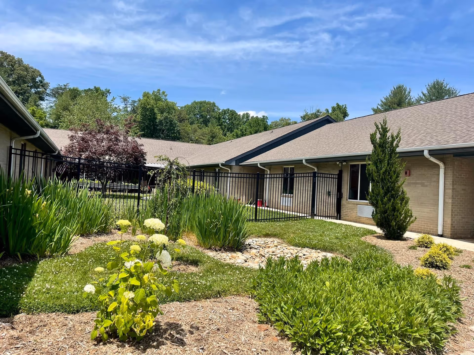 Outdoor garden area at Swannanoa Valley Health and Rehabilitation with various green plants, shrubs, and a small rock bed. The garden is enclosed by a black metal fence, and the building with beige brick walls and a gray roof is visible in the background under a partly cloudy blue sky.
