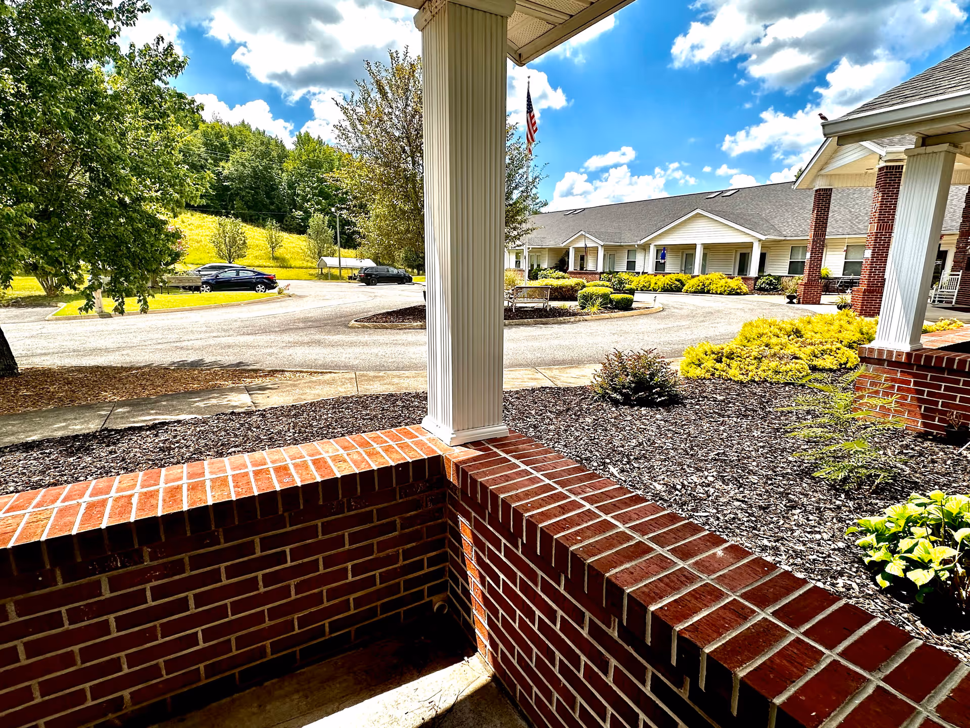 View from a covered porch area with red brick walls and white columns looking out onto a circular driveway, landscaped bushes, trees, and a single-story building under a partly cloudy blue sky.