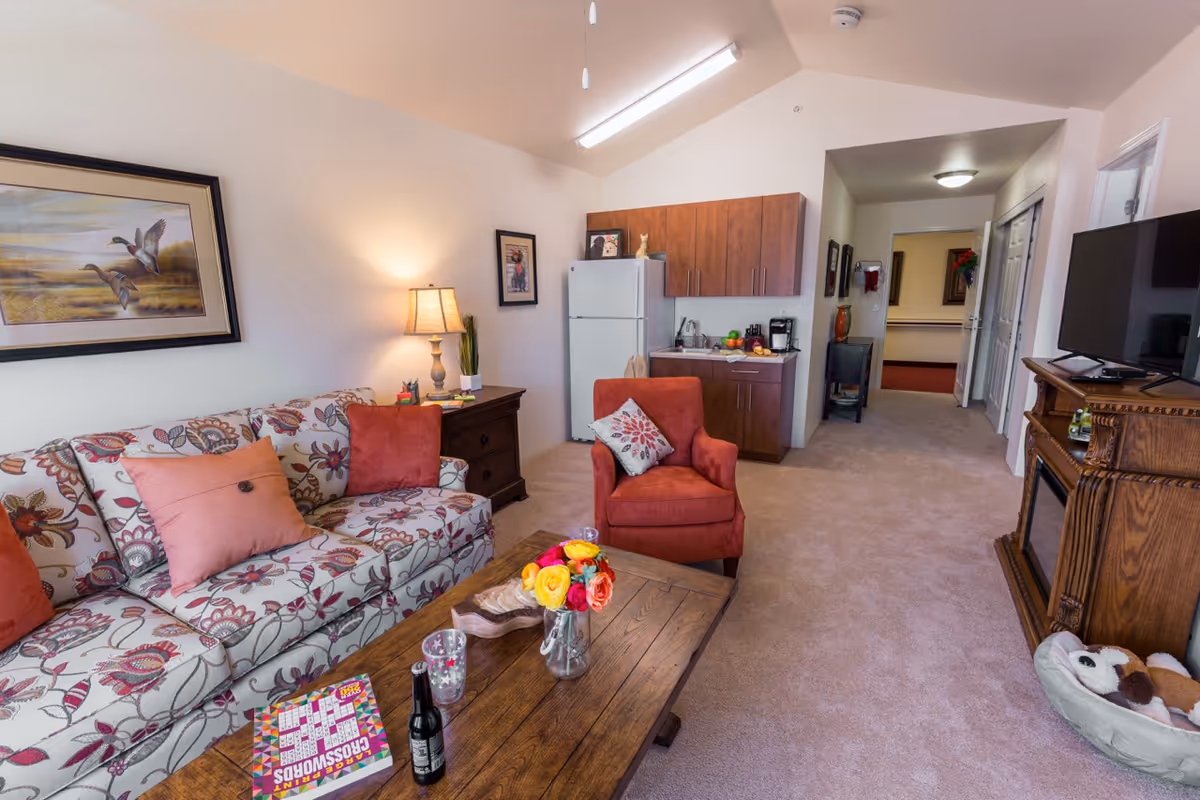 A cozy living room area in a senior living facility featuring a floral patterned sofa with orange and beige cushions, an orange armchair with a decorative pillow, a wooden coffee table with a vase of colorful flowers, a crossword puzzle book, and a bottle. The room also includes a wooden side table with a lamp, a small kitchenette with a refrigerator, cabinets, and a coffee maker, and a wooden TV stand with a flat-screen television. The carpeted floor and neutral wall colors create a warm and inviting atmosphere.