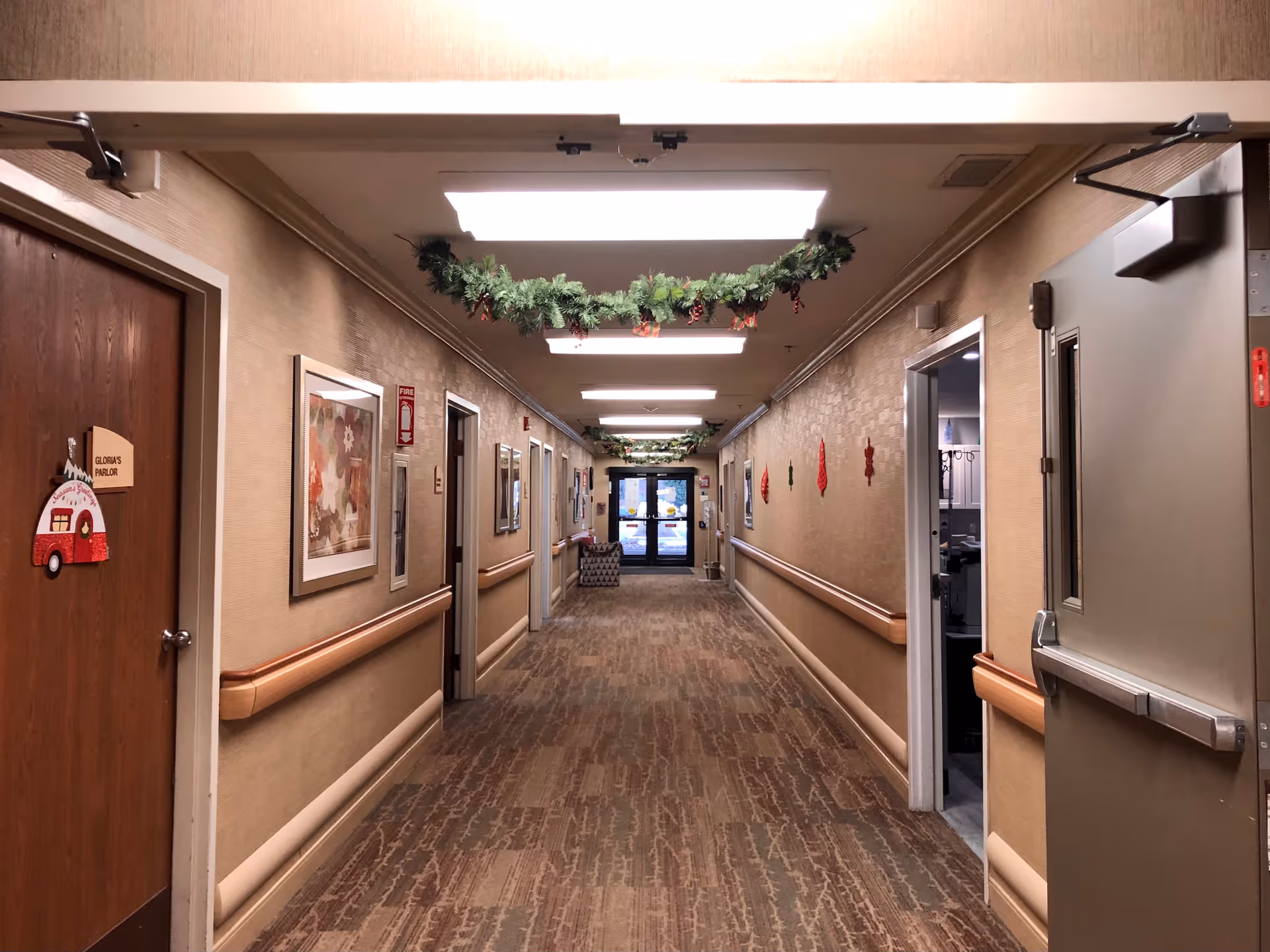 A long, well-lit hallway in a senior living facility decorated with green garlands hanging from the ceiling. The hallway has beige walls with handrails on both sides, framed artwork, and several doors, including one labeled 'Gloria's Parlor'. The floor is carpeted with a patterned design, and there is a glass door at the far end of the hallway.