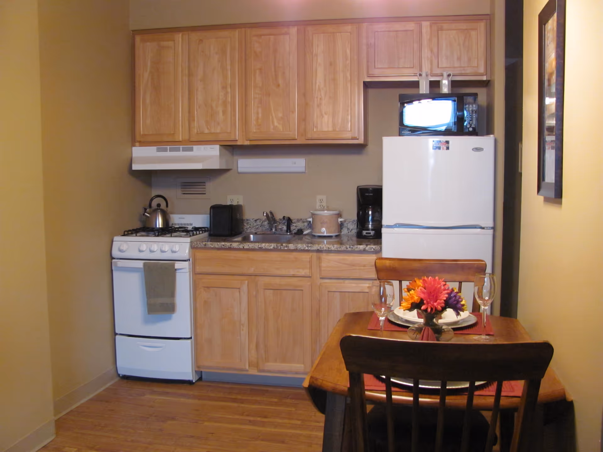 Small kitchen area with wooden cabinets, a white stove with a kettle, a toaster, a slow cooker, a coffee maker, and a white refrigerator with a microwave on top. In front of the kitchen is a wooden dining table set for two with wine glasses and a small vase of colorful flowers.