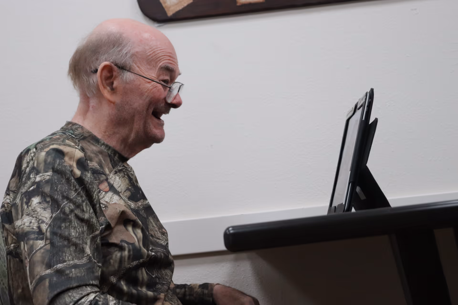 An elderly man wearing glasses and a camouflage-patterned shirt is sitting at a table, smiling and looking at a tablet device propped up in front of him against a plain white wall.