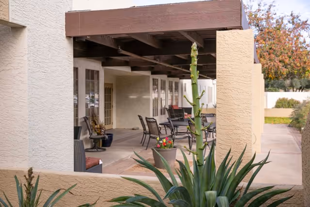 Outdoor covered patio area with several black metal chairs and tables, beige stucco walls, and a large green plant in the foreground. The patio is part of a building with multiple glass doors and windows, and there are trees and shrubs visible in the background.
