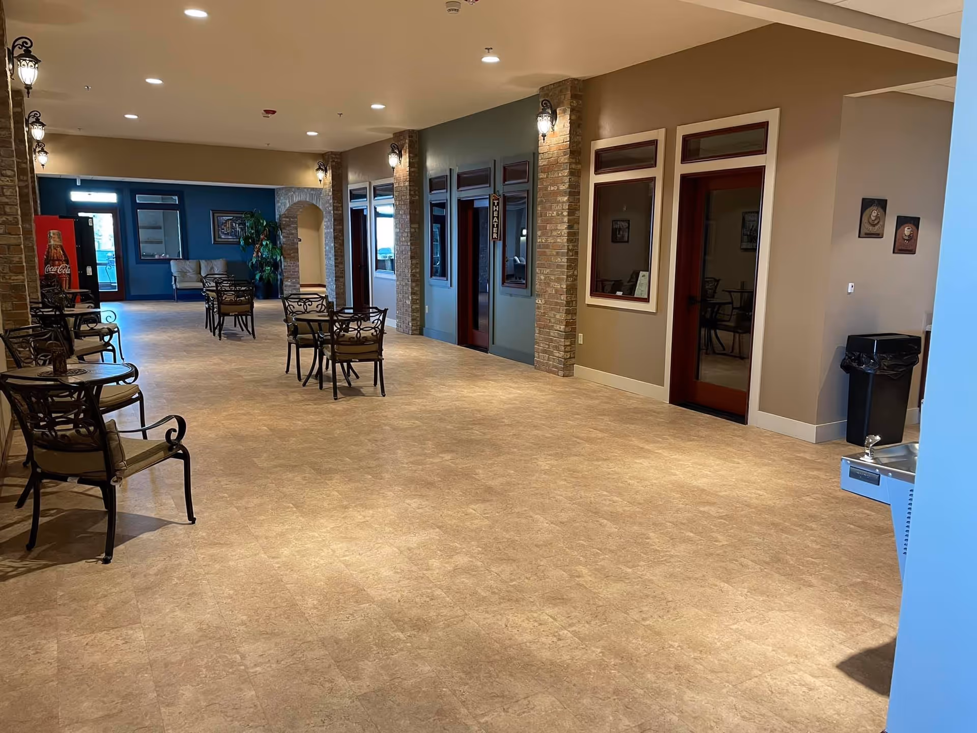 Interior hallway of an assisted living facility with beige tiled flooring, brick columns, and wall-mounted lantern-style lights. Several small tables with chairs are arranged along the hallway. There is a water fountain on the right side and a Coca-Cola vending machine in the background near a blue wall with a window and a door. The hallway leads to a room labeled 'Theater'.