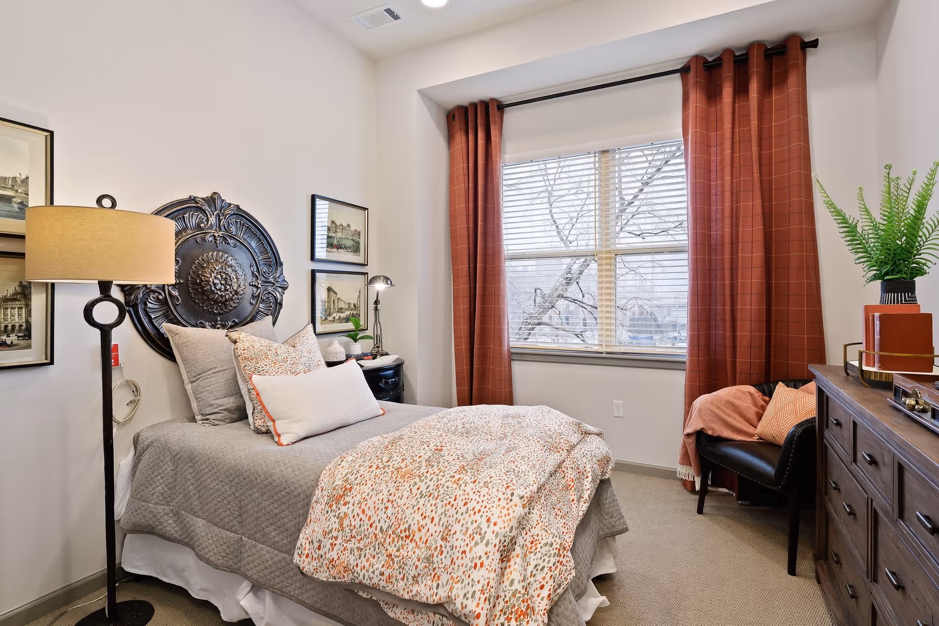 A cozy bedroom with a single bed dressed in gray and patterned bedding, a floor lamp beside the bed, framed artwork on the wall, a window with red curtains, a black chair with a pink throw blanket and pillow, and a wooden dresser topped with a green plant and decorative items.