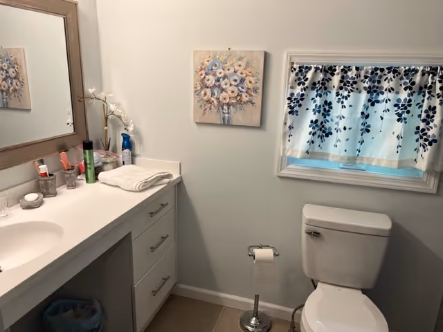 A clean bathroom with a white countertop and sink on the left, a large mirror above the sink, and various toiletries on the counter. A white towel is folded on the counter. On the right side, there is a white toilet with a toilet paper holder next to it. Above the toilet is a window with a white frame and a blue and white floral curtain. A floral painting hangs on the wall between the mirror and the window.