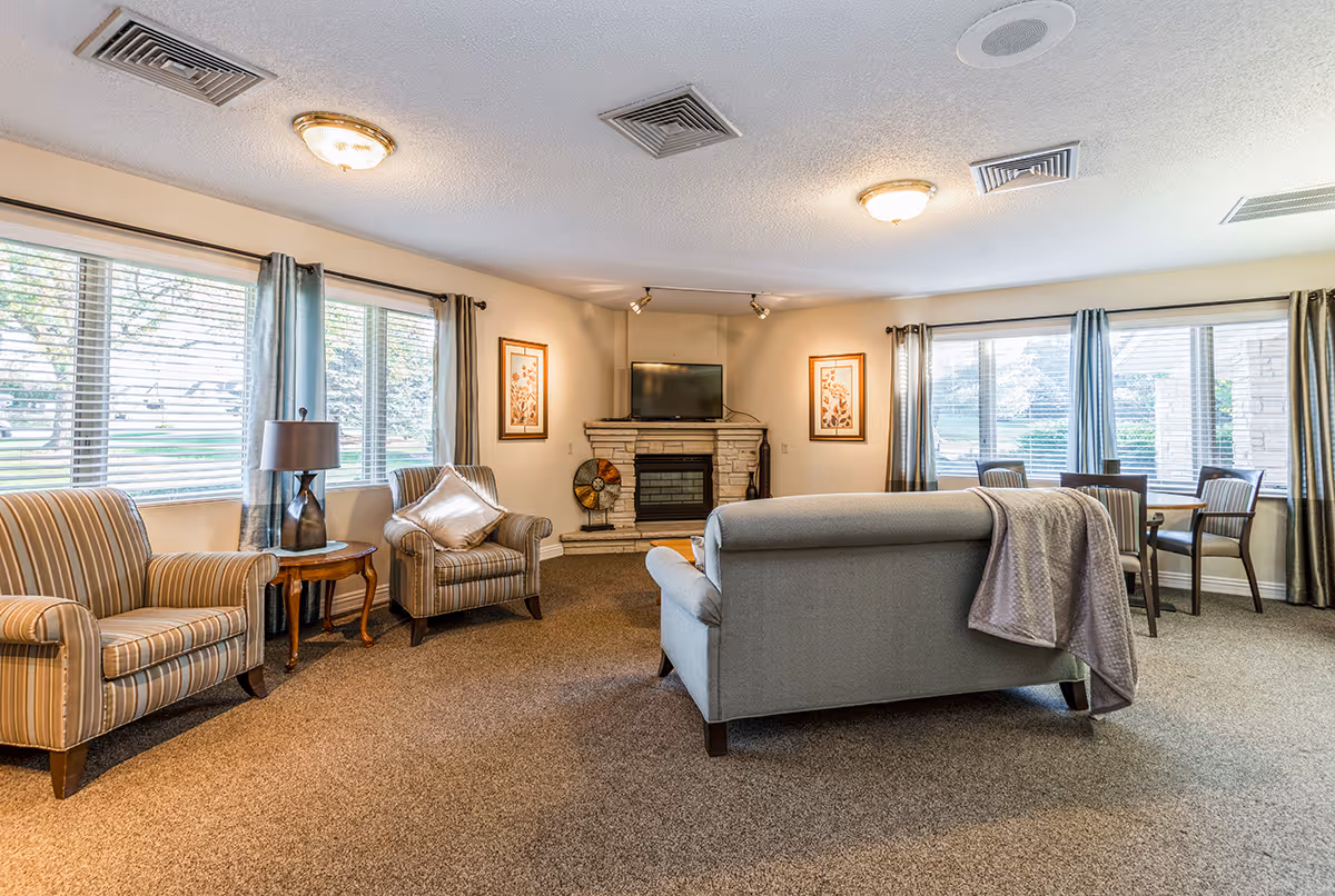 A cozy senior living common area with a gray couch, two striped armchairs, a wooden side table with a lamp, a stone fireplace with a TV mounted above it, and a small dining table with four chairs near large windows with curtains.