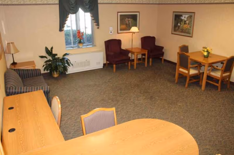 A common area in a senior living facility featuring a striped sofa, two burgundy armchairs with a wooden side table and lamp between them, a wooden dining table with four chairs, a potted plant near a window with green curtains, and framed artwork on the walls.