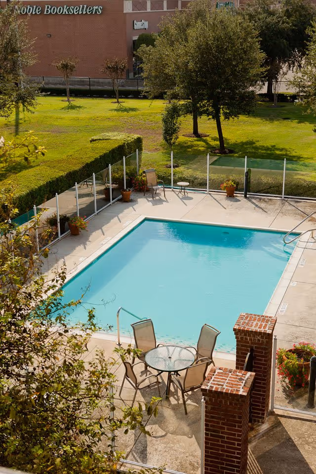 Outdoor swimming pool area with clear blue water, surrounded by a concrete deck with a glass table and four chairs. There are potted plants and a safety fence around the pool. In the background, there is a grassy area with trees and a building with a sign that reads 'Noble Booksellers'.