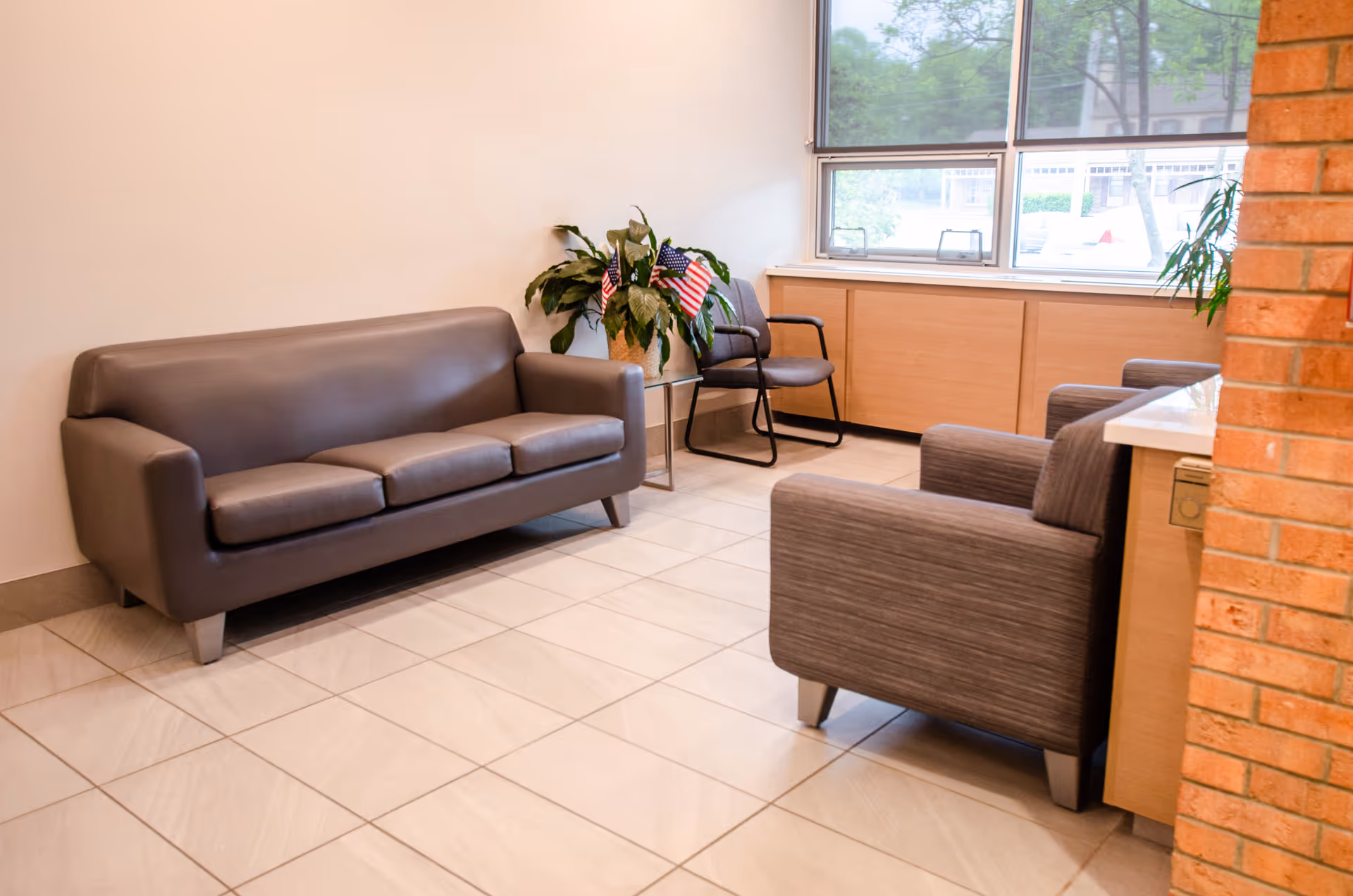 A waiting area with a gray leather sofa, two gray armchairs, and a gray chair with armrests near a window. There is a potted plant with small American flags on a side table next to the sofa. The floor is tiled, and there is a brick column on the right side.