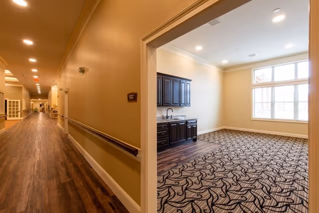 View down a long hallway with wood flooring and beige walls, featuring handrails on the right side. To the right, there is an open room with patterned carpet, a large window letting in natural light, and dark wood cabinets with a countertop and sink.