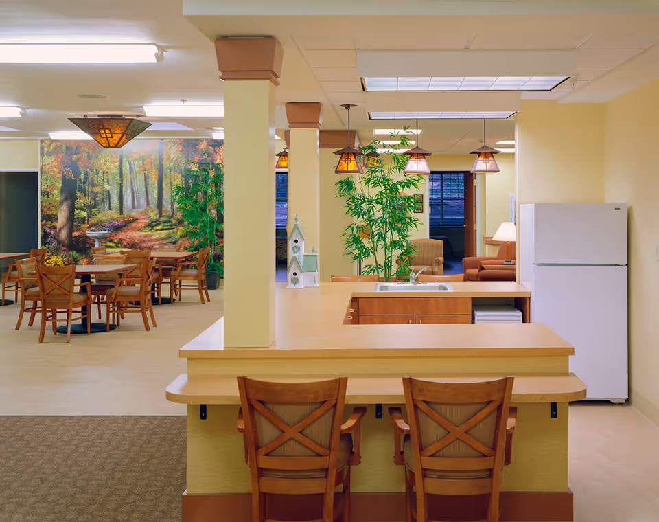 Interior view of a senior living facility showing a kitchen area with a countertop and two wooden chairs in the foreground. Behind the kitchen is a dining area with several wooden tables and chairs, and a large wall mural depicting a forest scene. There are hanging pendant lights above the kitchen counter and green plants near the back. In the background, a living room area with armchairs and a lamp is visible.