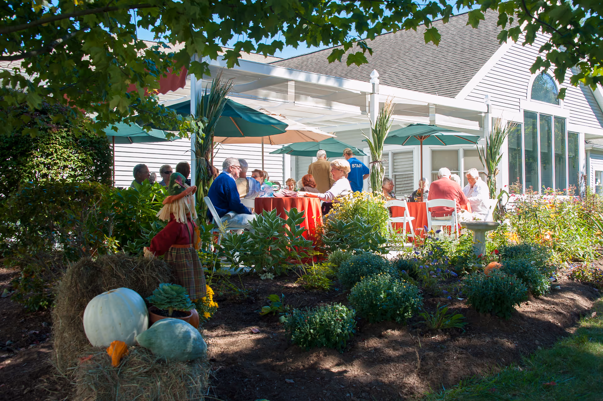 A group of elderly people sitting and socializing at tables with red tablecloths under umbrellas on a patio outside a white building. The patio is surrounded by a garden with green plants, flowers, hay bales, and decorative pumpkins. A scarecrow decoration is also visible near the hay bales.