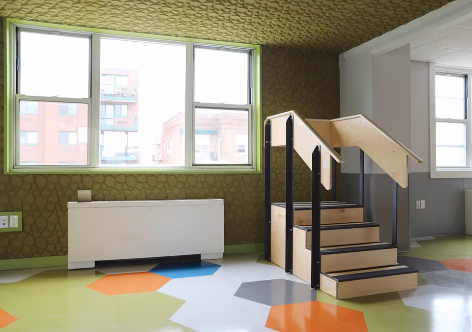 A brightly lit room with large windows, green and textured walls, a white radiator cover, colorful hexagonal floor tiles in orange, blue, gray, and green, and a small wooden staircase with handrails on both sides.