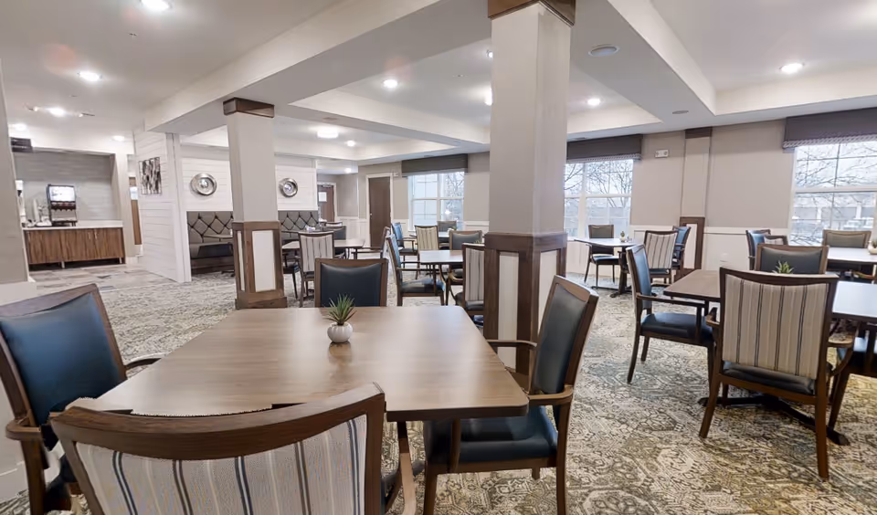 Bright dining room in a senior living facility with multiple wooden tables and upholstered chairs, pillars, and large windows.