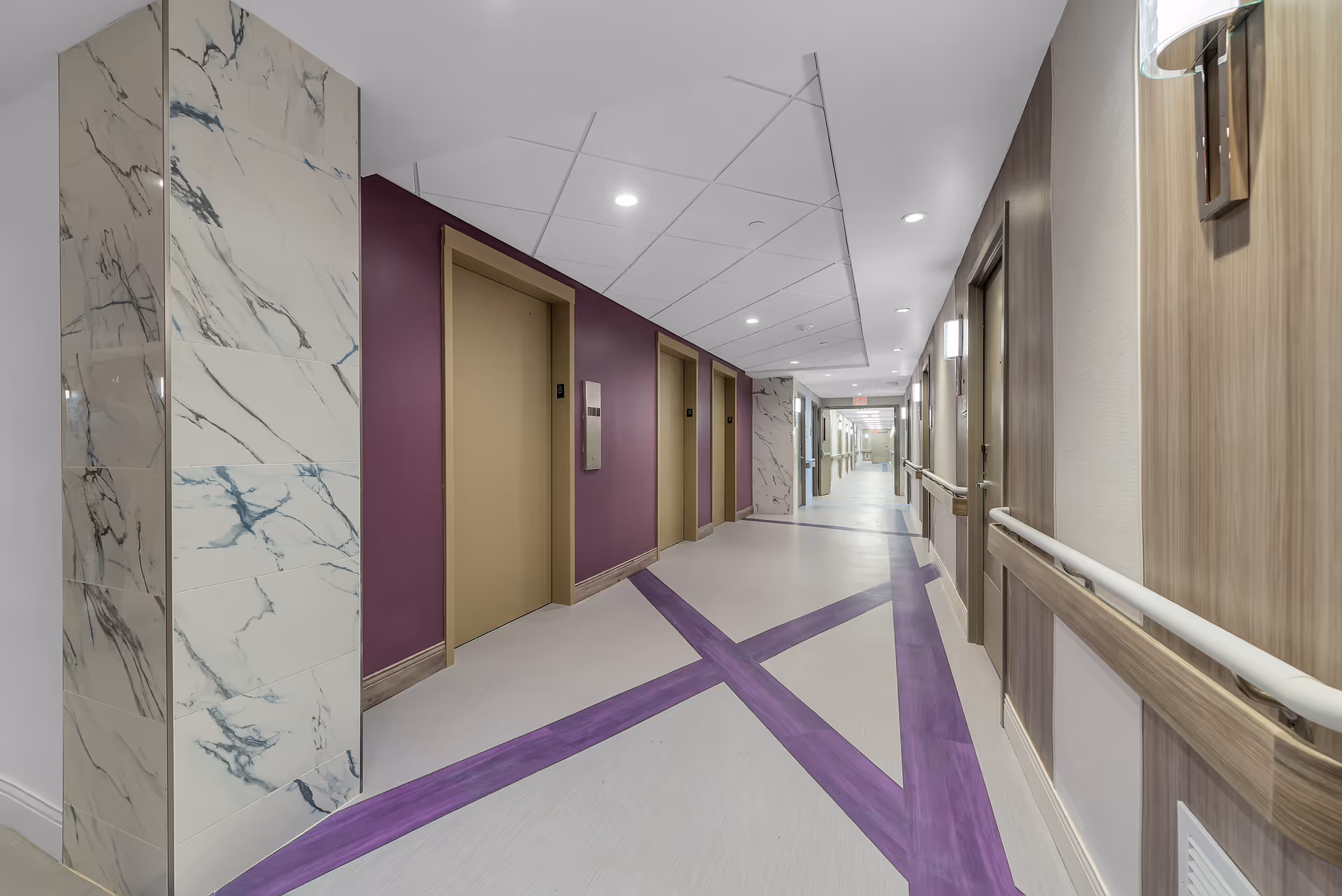 A clean, modern hallway in an assisted living facility with beige doors, purple accent walls, and marble-patterned columns. The floor features a unique design with intersecting purple lines. Handrails run along the walls, and ceiling lights illuminate the corridor.