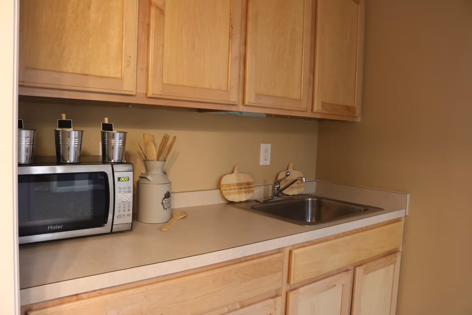 A small kitchen area with light wood cabinets, a countertop with a stainless steel sink, a microwave, and kitchen utensils in a ceramic container. Two wooden cutting boards shaped like pumpkins are placed against the wall behind the sink.