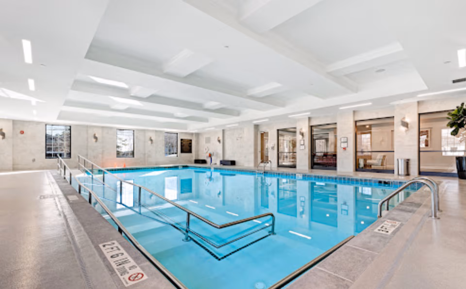 Bright indoor swimming pool with handrails, seating areas and large windows inside the facility.
