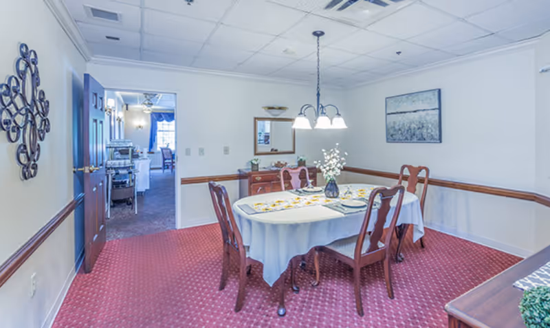 A dining room with a round table covered with a white tablecloth, set with plates and a vase of flowers in the center. Four wooden chairs surround the table. The room has red carpet flooring, white walls with a wooden chair rail, a wall mirror, a painting, and a decorative wall piece. A doorway leads to another room with more seating and a window.