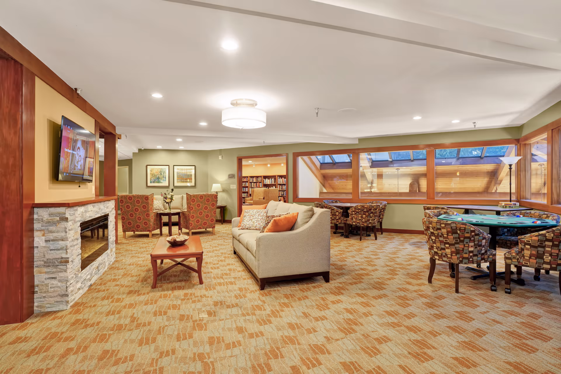 A spacious and well-lit common area in a senior living facility featuring a stone fireplace with a mounted flat-screen TV, a beige sofa with decorative pillows, two patterned armchairs around a small table, and additional seating with patterned chairs around tables near large windows. The room has warm lighting, carpeted floors, and a cozy, inviting atmosphere.
