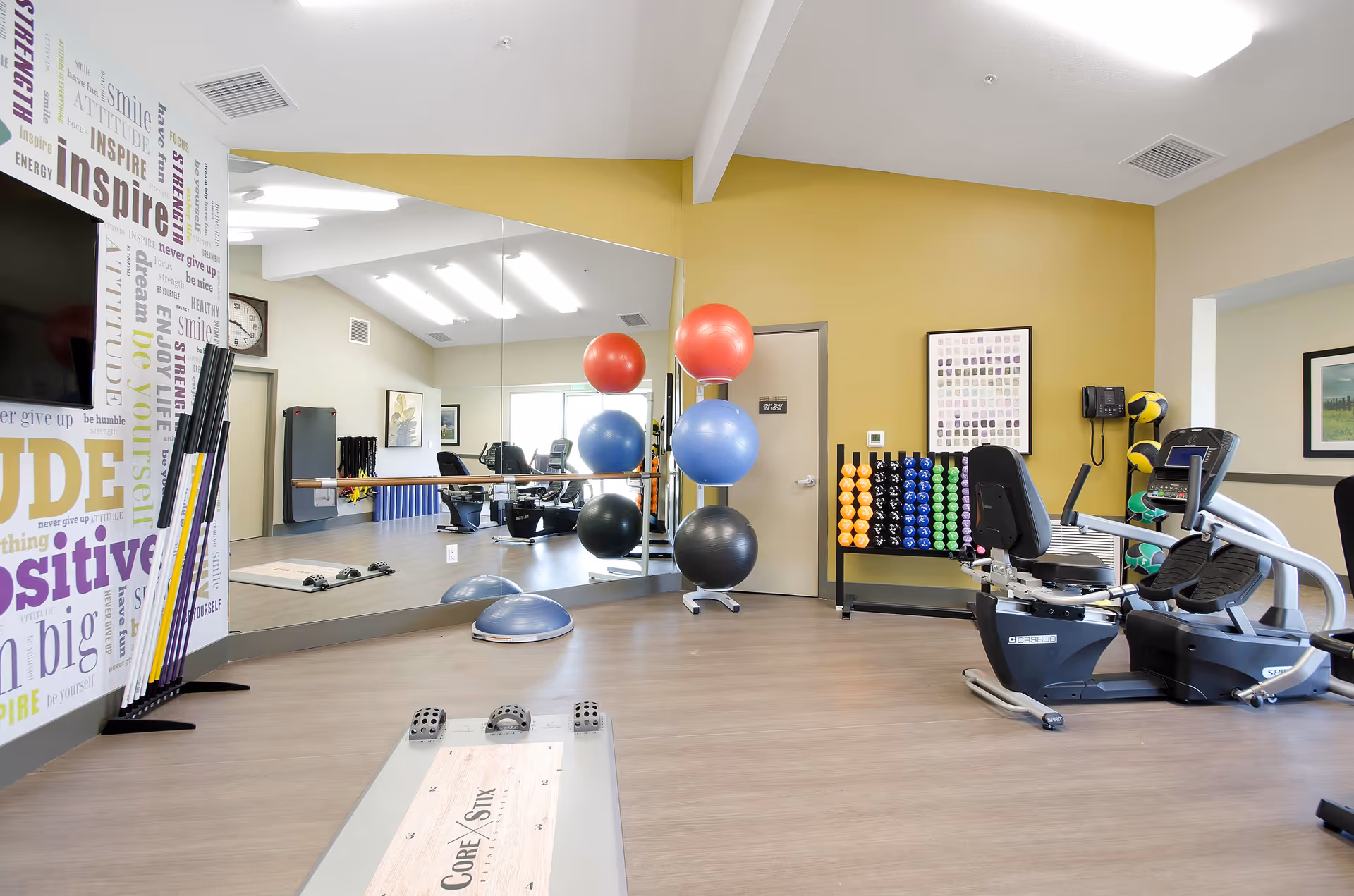 A bright and spacious fitness room in an assisted living facility featuring exercise equipment including a recumbent bike, colorful exercise balls stacked on a rack, a variety of dumbbells organized on a rack, and a mirrored wall reflecting the room. The walls are painted yellow and beige, with motivational words displayed on one wall. The floor is wood-style, and the room has ample lighting from ceiling fixtures.