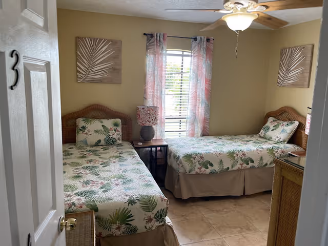 A bedroom with two twin beds featuring floral and leaf-patterned bedding. Each bed has a matching pillow and a wicker headboard. Between the beds is a small wooden nightstand with a decorative lamp. The room has beige walls, a window with pink and white curtains, and two framed leaf artworks hanging above each bed. The floor is tiled, and a ceiling fan with a light is visible.