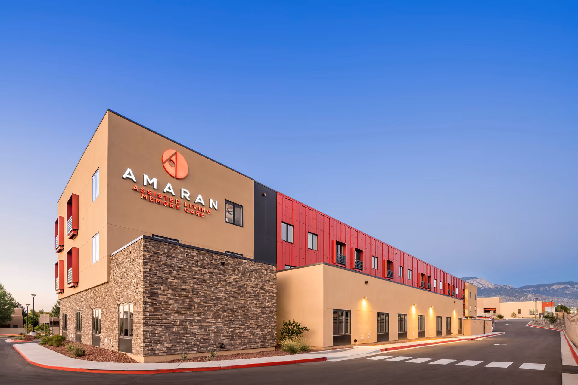 Exterior view of Amaran Senior Living facility building during dusk, featuring a modern design with beige, stone, and red paneling, clear sky, and a paved road with a crosswalk in front.