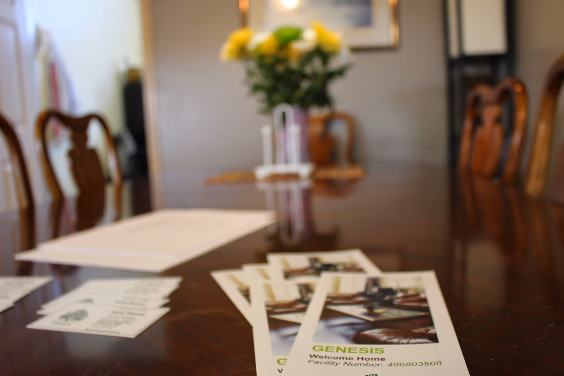 A polished wooden dining table with brochures and papers spread out on it. In the background, there are wooden chairs and a vase with yellow flowers on the table.