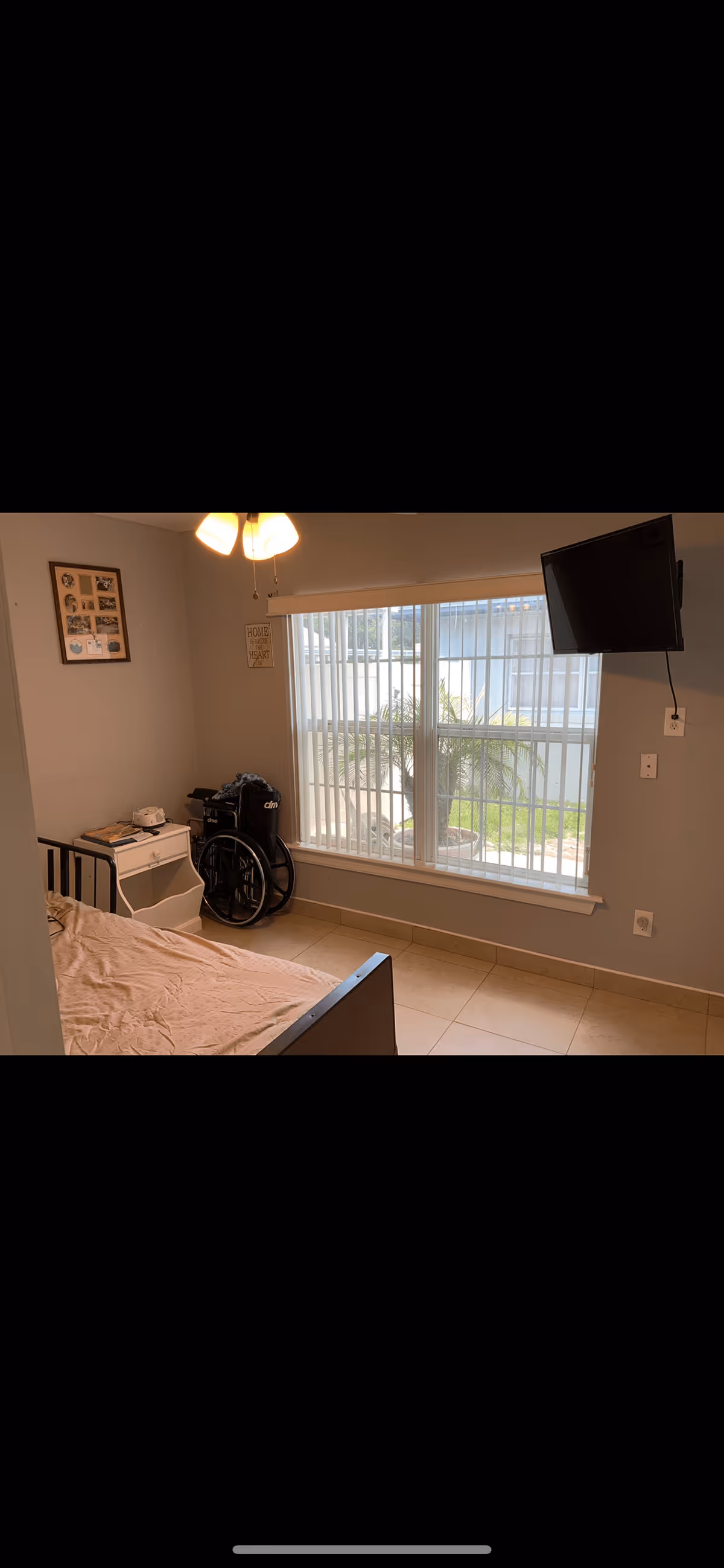 Small bedroom with a bed, bedside table, wheelchair, wall-mounted TV, and a window with vertical blinds overlooking a courtyard.