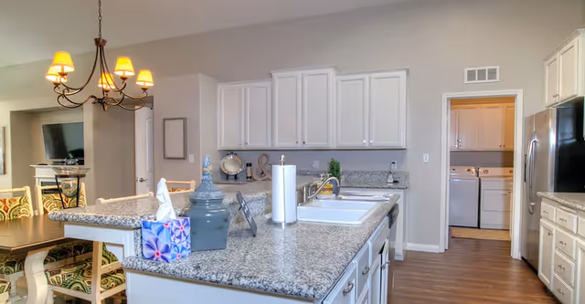 Open kitchen with a large granite island, white cabinetry, stainless refrigerator, sink, and a view into the dining area and laundry room.