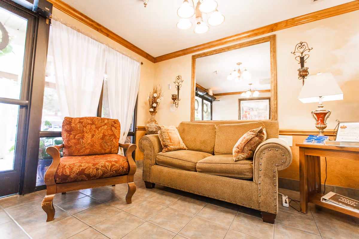 Warmly lit seating area with a patterned armchair and a cushioned sofa, a large mirror, and a side table in a reception-like room.