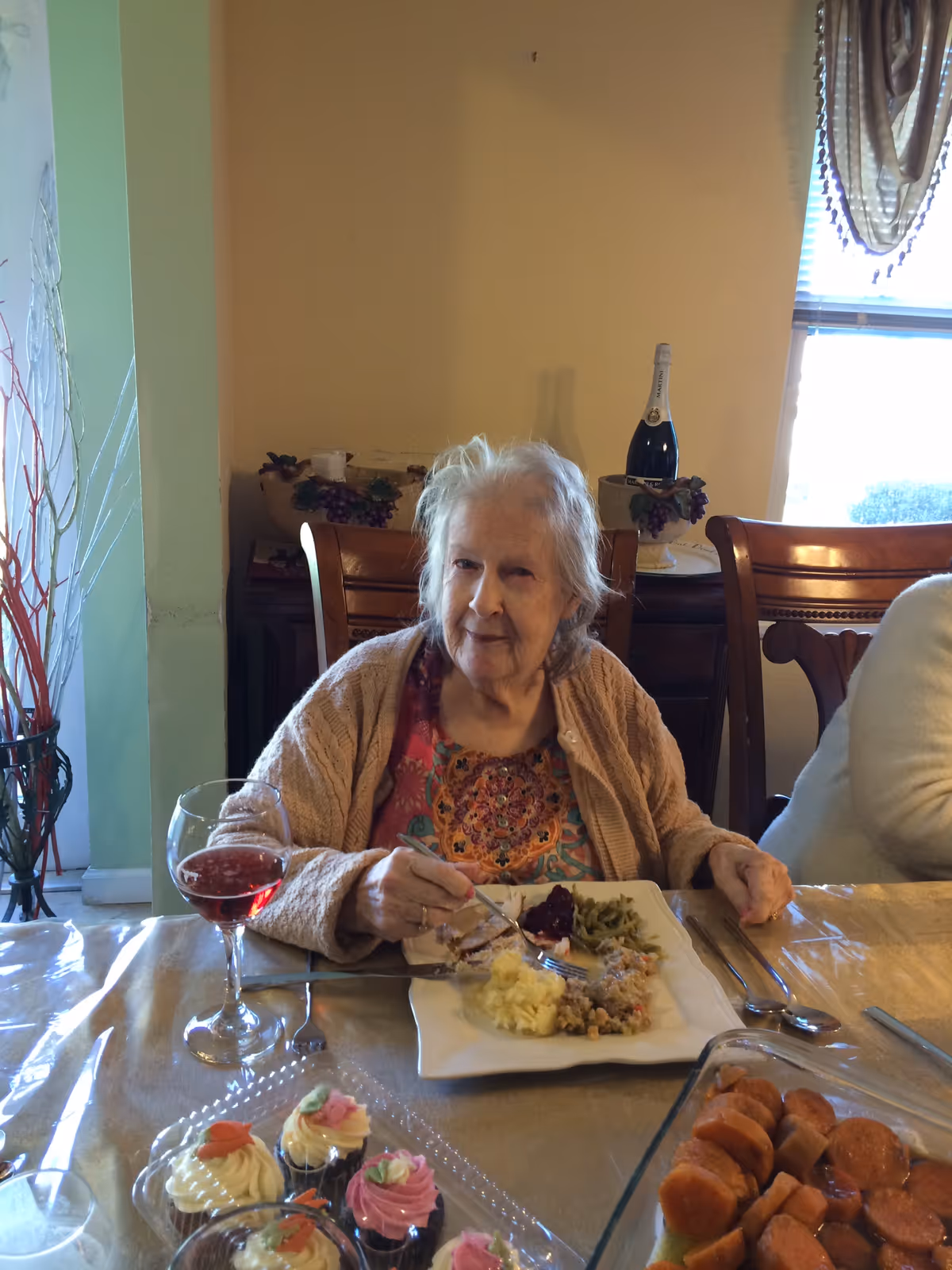An elderly woman sitting at a dining table with a plate of food in front of her, including mashed potatoes, green beans, and cranberry sauce. She is wearing a colorful top and a beige cardigan, holding a fork. There is a glass of red beverage and a tray of decorated cupcakes on the table. The background shows wooden chairs, a window with curtains, and a bottle on a side table.