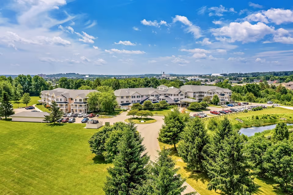 Aerial view of Solstice Senior Living at Bangor, showing a large multi-story building surrounded by green lawns, trees, and a small pond under a partly cloudy blue sky. Several cars are parked in the parking lots adjacent to the building.