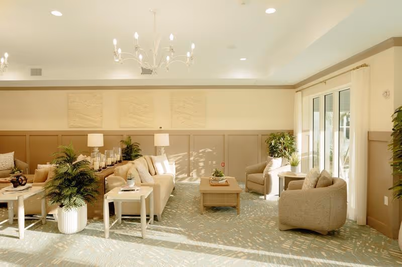 A bright and spacious living room area with beige walls and carpeted floor. The room features comfortable beige sofas and armchairs arranged around a wooden coffee table. There are side tables with lamps and decorative plants placed throughout the room. Large windows with sheer white curtains allow natural light to fill the space.