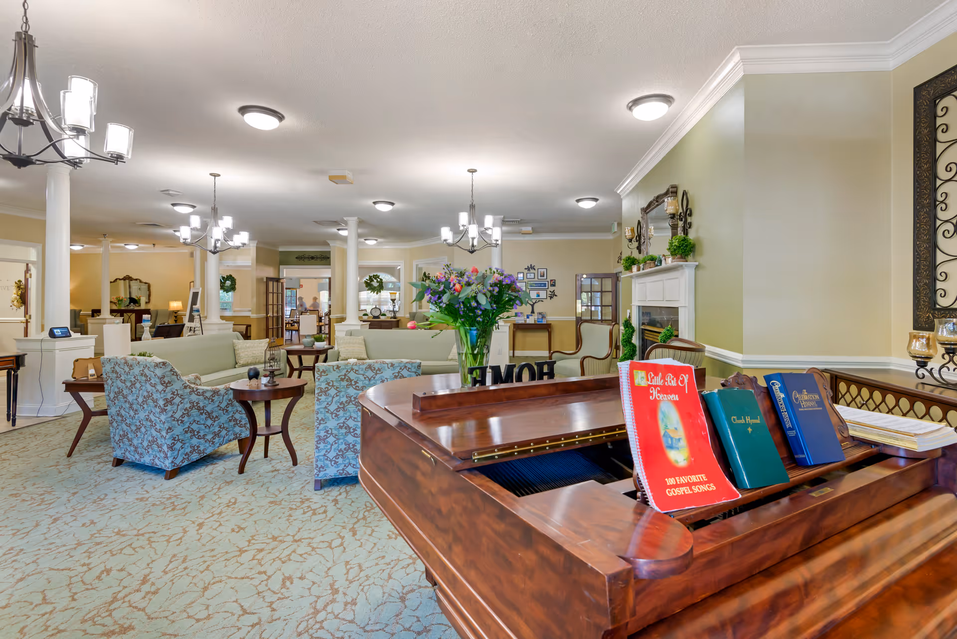 A spacious and well-lit living room area in a senior living facility with comfortable seating including sofas and armchairs arranged around small tables. A wooden piano with hymn books and a vase of flowers on top is in the foreground. The room features light-colored walls, carpeted floors, chandeliers, and decorative elements such as framed pictures and plants.