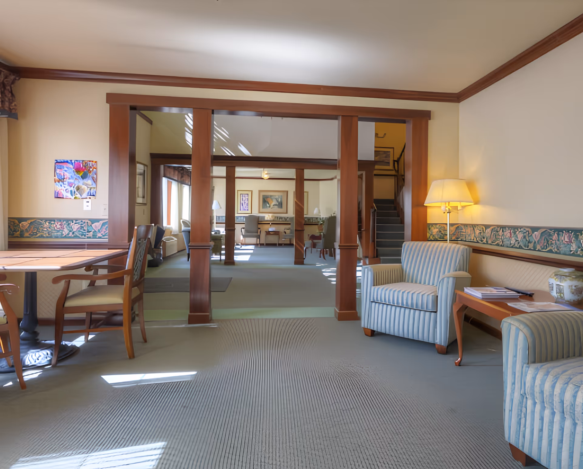 A spacious and well-lit common area in a senior living facility featuring comfortable striped armchairs, a wooden side table with books and a lamp, and a wooden dining table with chairs. The room has beige walls with decorative border trim, large wooden columns dividing the space, and carpeted flooring. Sunlight streams in through windows, creating a warm and inviting atmosphere.