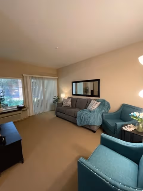 A cozy living room with a gray sofa adorned with patterned pillows and a teal throw blanket. Two teal armchairs with nailhead trim face the sofa, with a small round black side table between them holding a vase of white flowers and a magazine. A large rectangular mirror hangs on the beige wall above the sofa. Vertical blinds cover a sliding glass door, and a window with blinds is visible on the left side. The room is softly lit by a floor lamp and a table lamp.