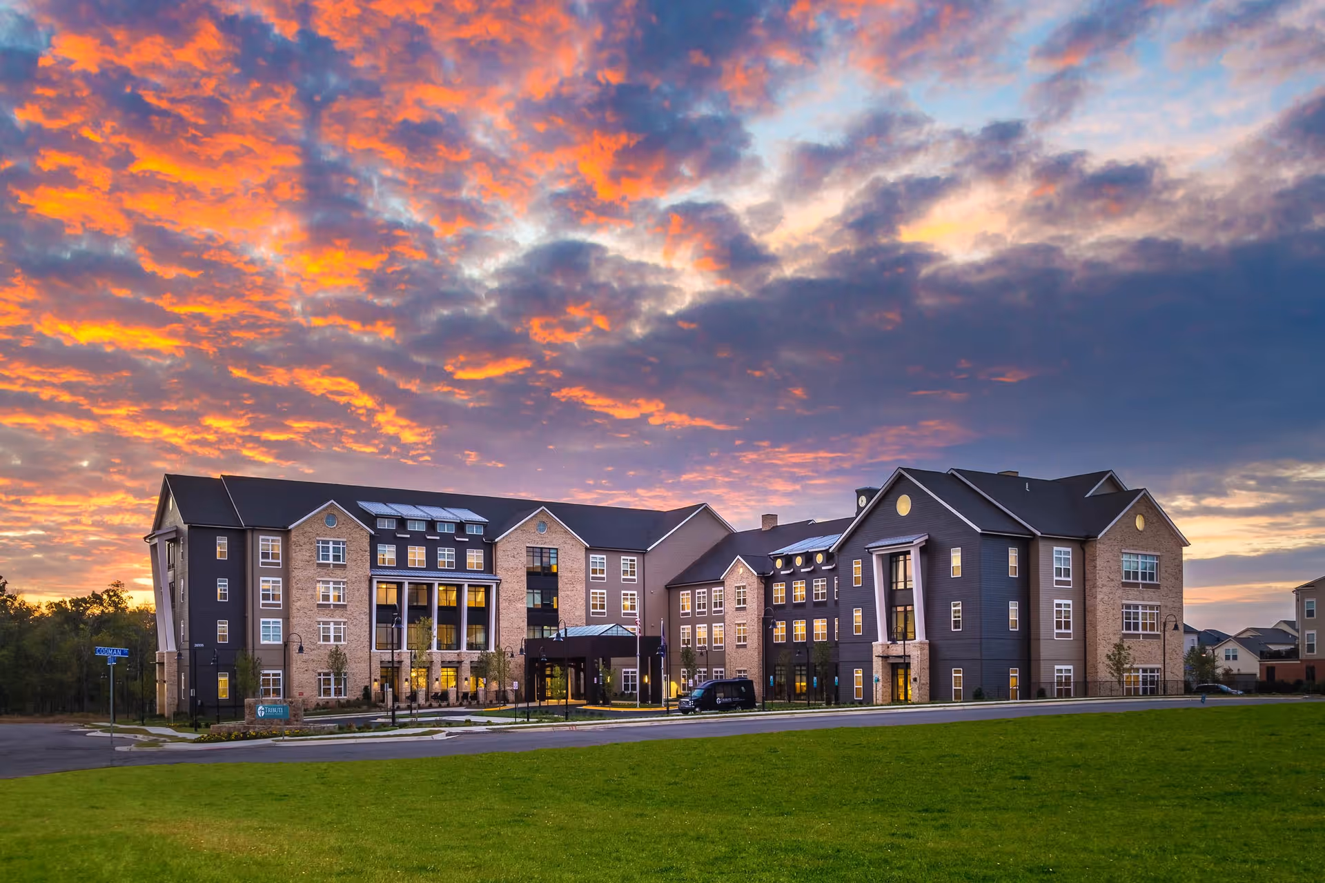 Exterior view of Tribute at One Loudoun senior living facility at sunset, showing a large multi-story building with a mix of brick and dark siding, numerous windows, and a covered entrance. The sky is filled with vibrant orange and purple clouds, and there is a green lawn in the foreground.