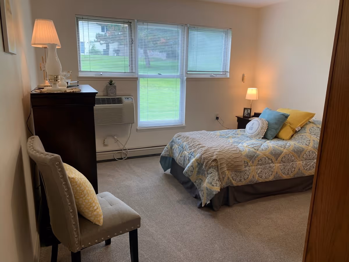 A cozy bedroom with a bed covered in a patterned quilt and several pillows, a nightstand with a small lamp and framed photo, a dark wooden dresser with a white lamp and decorative items, a beige upholstered chair with a yellow pillow, and three windows with blinds letting in natural light.