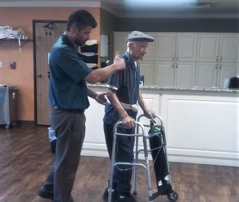 A caregiver giving a thumbs-up to an elderly man using a walker with an attached oxygen tank inside a room with wooden flooring and white cabinets in the background.