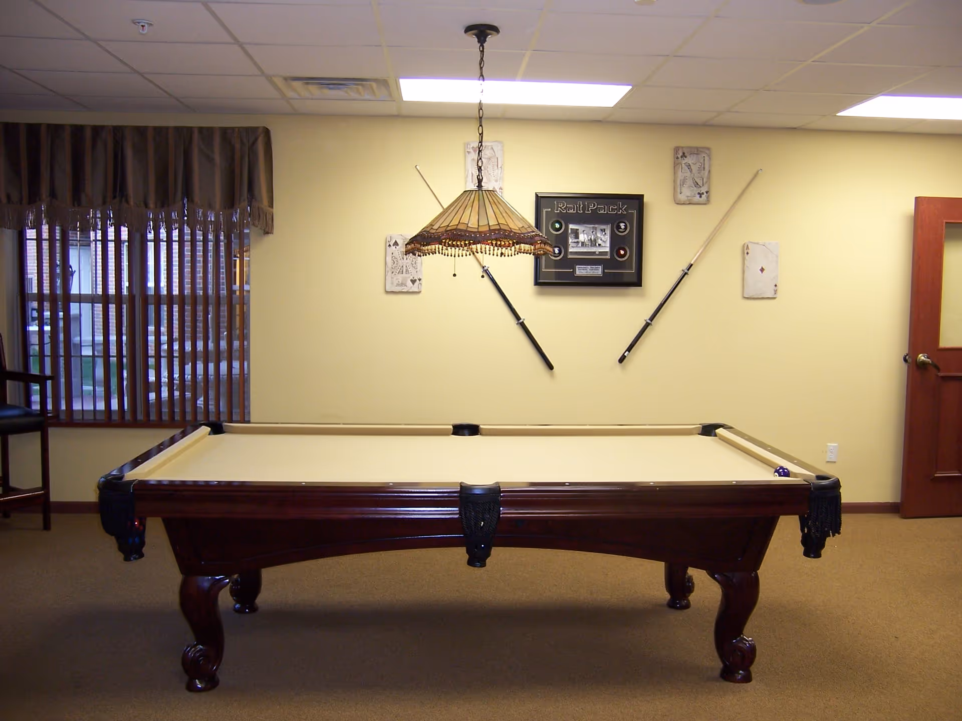A room with a beige pool table in the center, a hanging stained glass light fixture above it, and two pool cues mounted on the wall. The wall also features a framed Rat Pack memorabilia and playing card decorations. There is a window with vertical blinds and a brown valance on the left, and a wooden door on the right.