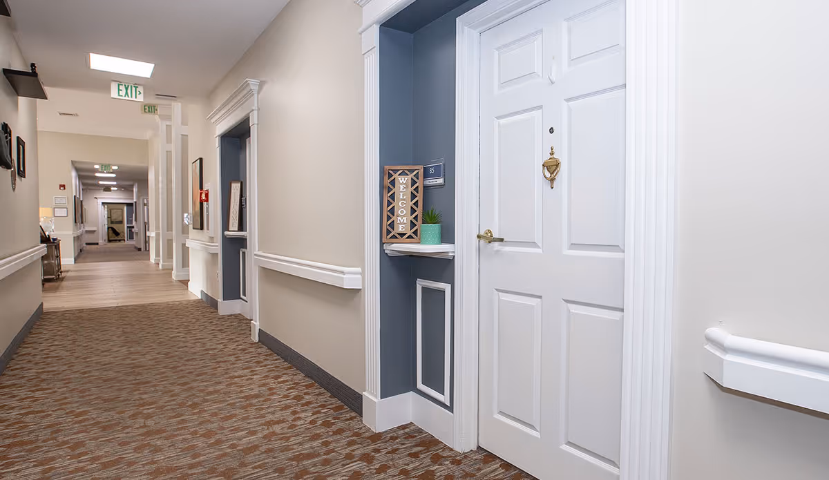 Carpeted corridor inside an assisted living facility showing a white apartment door with a welcome sign in a recessed alcove and handrails along the walls.