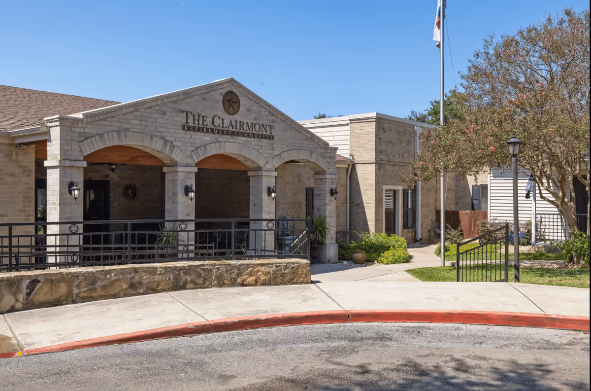 Exterior view of The Clairmont Retirement Community building with a covered entrance featuring stone arches, a ramp with black railings, a flagpole, and surrounding greenery under a clear blue sky.