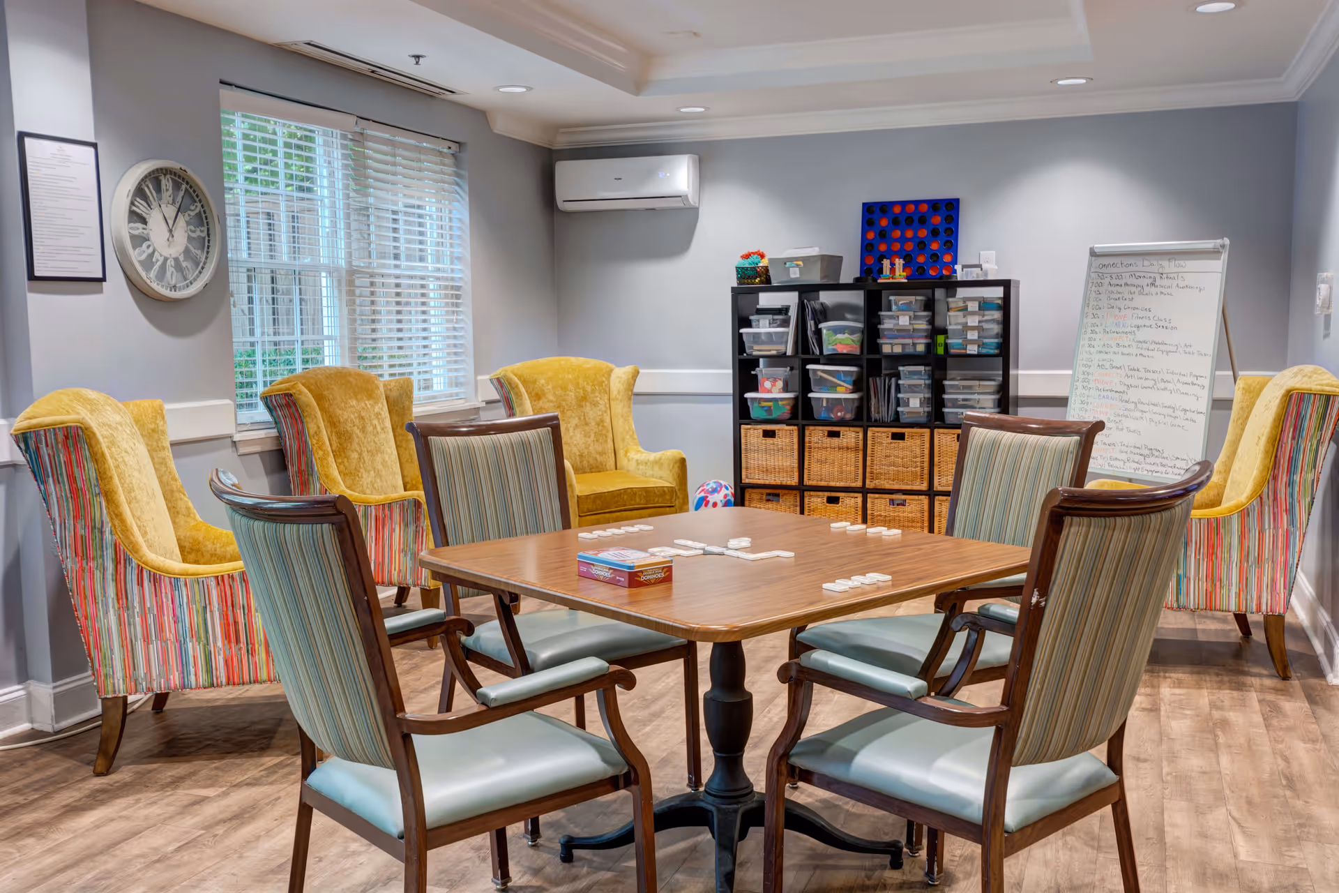 A cozy activity room with a wooden table and four chairs in the center, set up for a game with dominoes. Around the room are four colorful armchairs with yellow cushions and striped backs. A large window with white blinds lets in natural light. On the wall is a large clock and a framed document. A black shelving unit holds various plastic bins and games, and a whiteboard with a list of activities is visible in the corner. The room has light gray walls and wood flooring.
