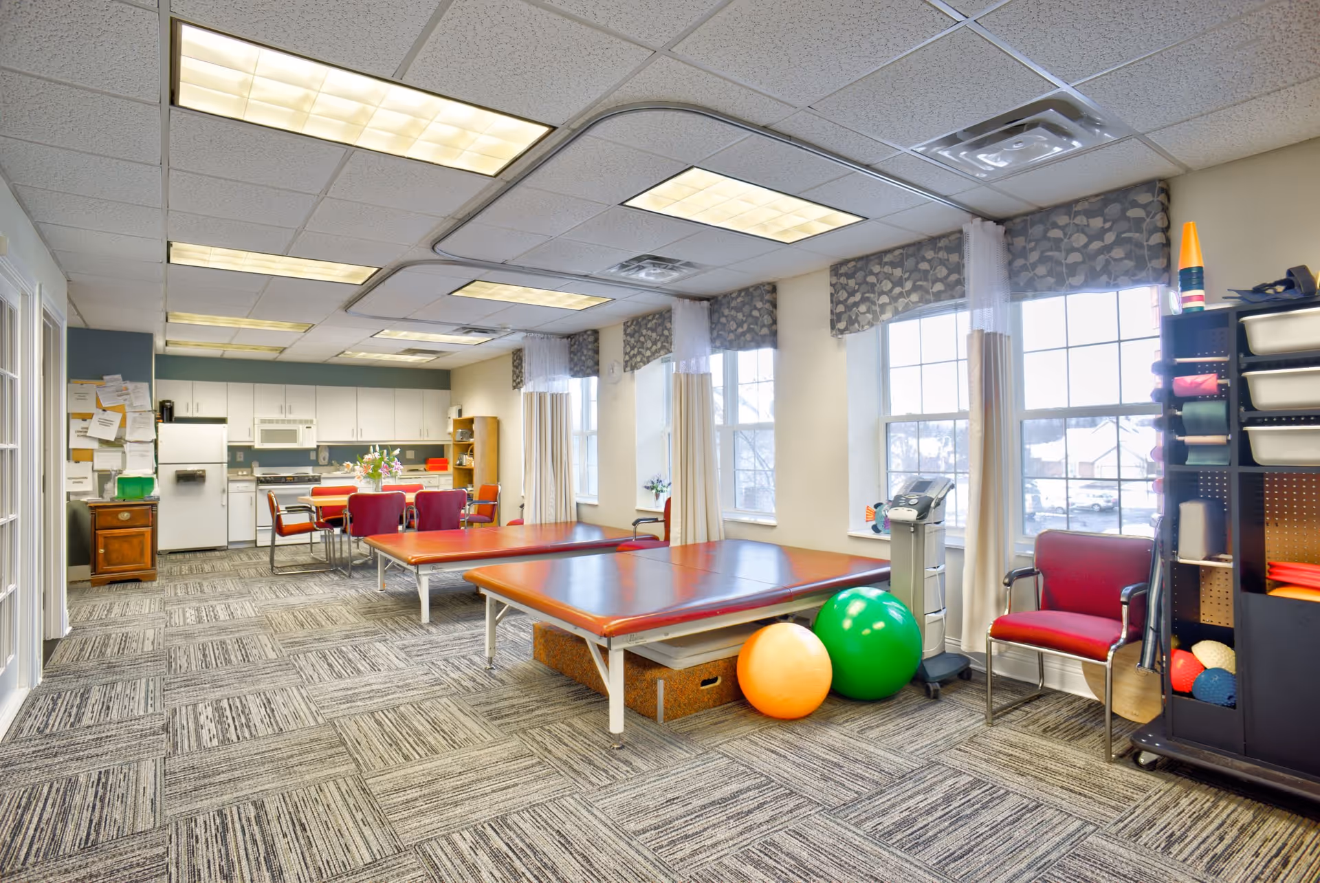 A bright therapy or activity room with two padded tables, exercise balls, a small kitchen area with white cabinets and appliances, several chairs, large windows with patterned valances and sheer curtains, and a shelving unit with various supplies.