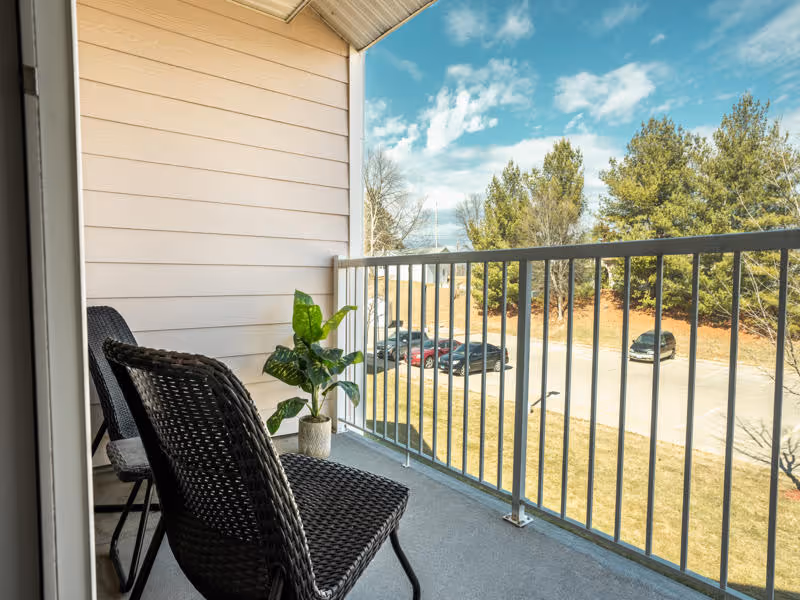 A small balcony with two black wicker chairs and a potted green plant. The balcony overlooks a parking lot with several cars and trees in the background under a partly cloudy sky.