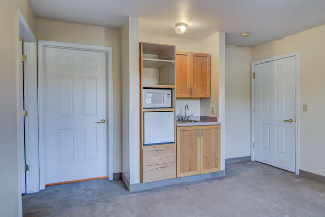 Small kitchenette area with wooden cabinets, a microwave, a mini refrigerator, and a sink in a room with beige walls and carpeted floor. Two white doors are visible on either side of the kitchenette.