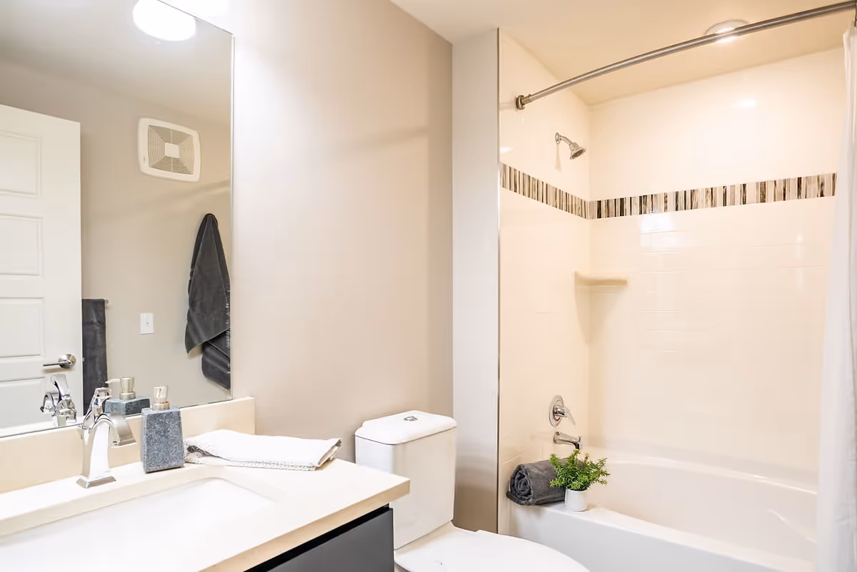 A clean and modern bathroom featuring a white sink with a silver faucet, a soap dispenser, and a folded towel on the countertop. There is a large mirror above the sink, a white toilet next to the sink, and a bathtub with a showerhead and a curved shower curtain rod. The bathtub area has white tiles with a decorative horizontal strip of darker tiles. A rolled dark gray towel and a small green plant are placed on the edge of the bathtub.