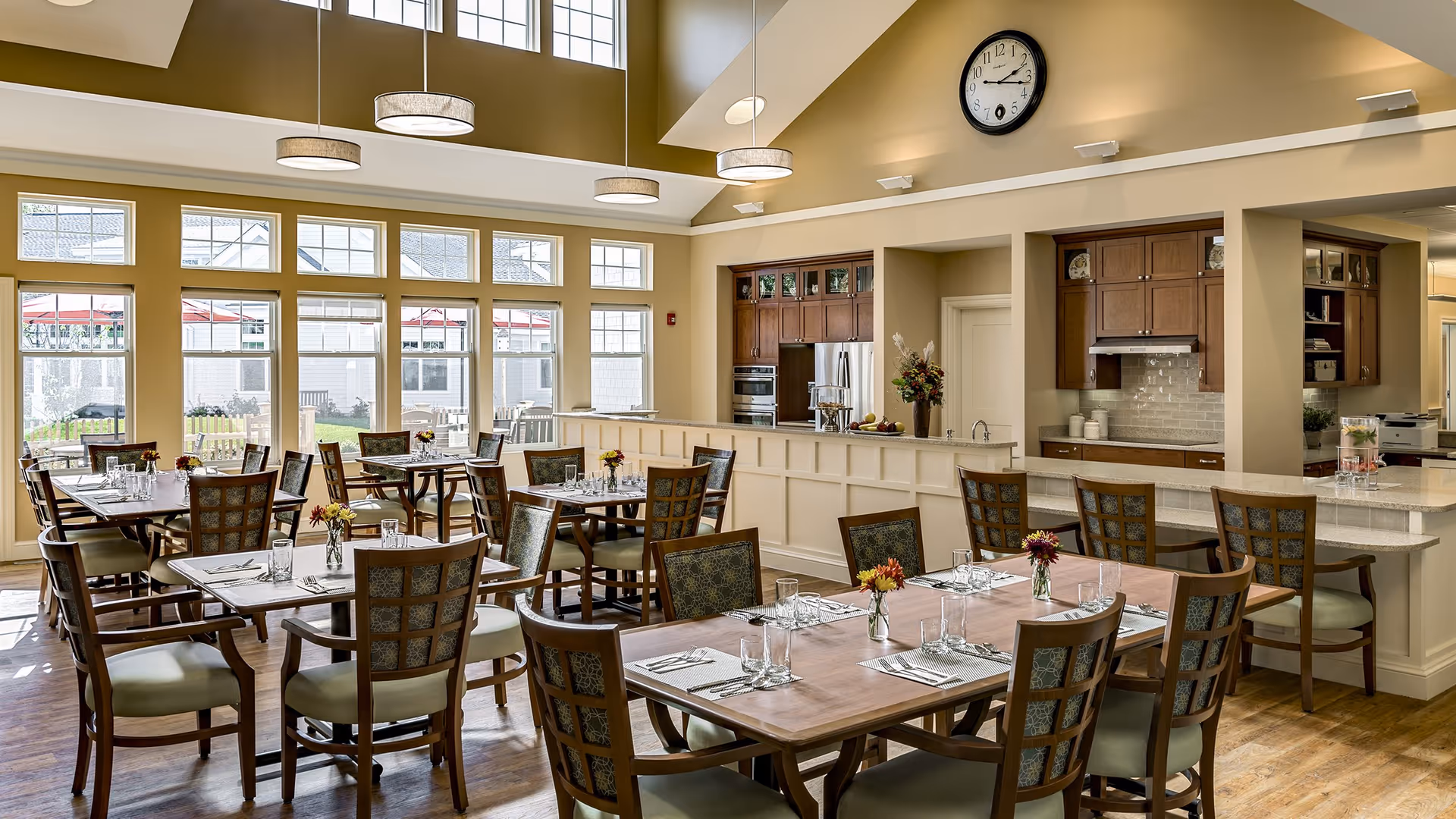 A bright dining room with multiple wooden tables and chairs arranged neatly. Each table is set with placemats, glasses, and small flower vases. Large windows allow natural light to fill the room, and a kitchen area with wooden cabinets and stainless steel appliances is visible in the background. A large clock is mounted on the beige wall above the kitchen area.